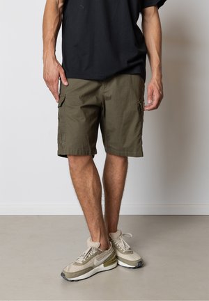 Mand iført olivengrøn cargo shorts, beige og hvide Nike sneakers og en sort t-shirt, stående mod en hvid væg.