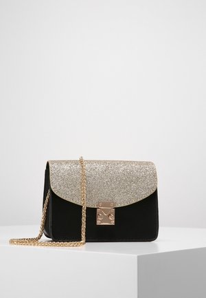 Bolso negro con solapa dorada brillante, cierre dorado y cadena dorada, apoyado sobre superficie blanca sobre un fondo liso.