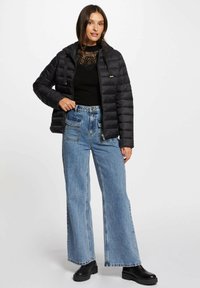 Giubbotto nero imbottito con cappuccio, chiusura con zip e dettagli dorati. Indossato sopra un top nero e abbinato a jeans in denim blu a gamba larga e stivali neri.