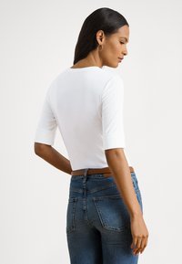 Sieviete ar gariem tumšiem matiem, valkājot baltu piegulošu kreklu ar elkoņa garuma piedurknēm un zilas džinsas ar brūnu jostu, skatās uz sāniem.