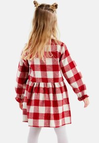 Punainen ja valkoinen ruudullinen mekko pitkillä hihoilla, röyhelöillä varustetuilla hihojen päissä ja kerätty vyötärö. Pehmeä flanellitekstuuri ja liehuva hame.