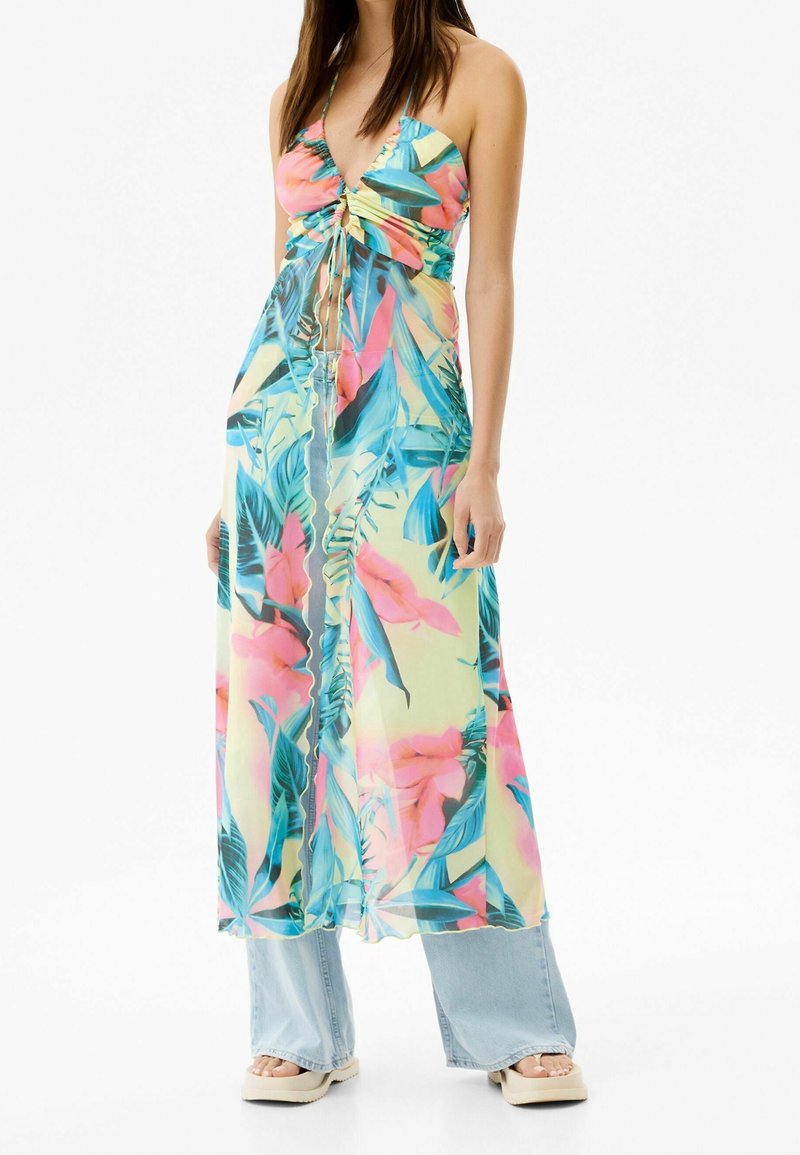 Vrouw in een kleurrijke tropical print halterjurk over lichtblauwe wijde jeans en crèmekleurige platform sandalen tegen een witte achtergrond.