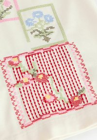 Tissu brodé présentant des motifs floraux en bleu, vert et rouge sur un fond crème. Montre un travail de points complexe et des textures variées.