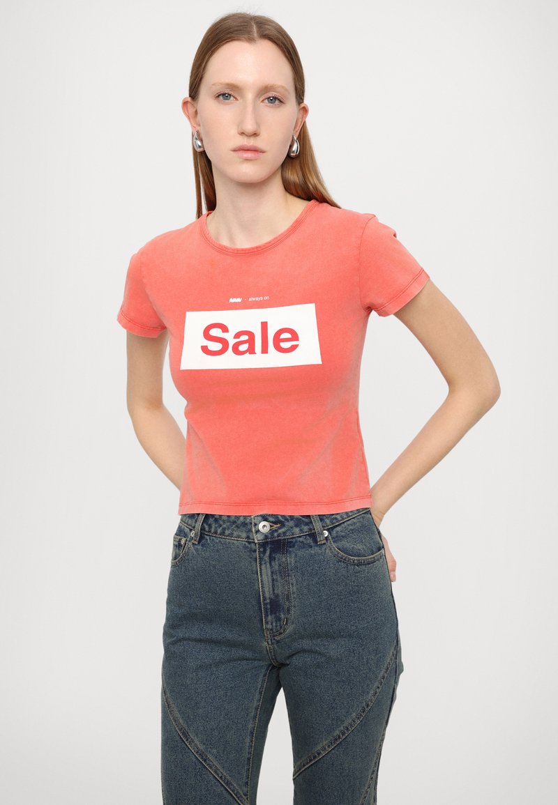 Camiseta corta de manga corta en color coral con un gran estampado blanco de "Venta", complementada con un corte relajado y una textura suave. Combinada con unos vaqueros de cintura alta.