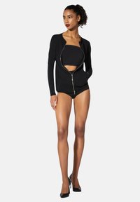 Svart zip-jacka med en rundad halsringning, figursydd design, långa ärmar och framfickor, bärs över en svart topp och shorts.