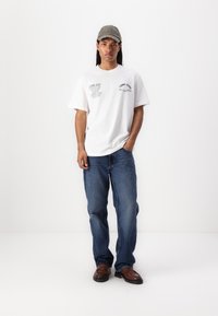 White short-sleeve t-shirt with graphic prints, paired with blue denim jeans. The model wears a gray cap and brown slip-on shoes.