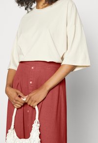 Personne portant un haut ample crème et une jupe rouge taille haute, tenant un sac à main tissé blanc avec les deux mains devant.