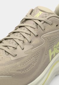 Beige running shoe with a textured mesh upper, contrasting yellow logo, and lacing system. Features a cushioned sole with a light accent.