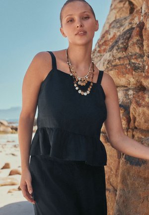 Jonge vrouw die een zwarte mouwloze top met ruches draagt en gelaagde gouden muntkettingen, staand bij een rotsachtig strand onder een heldere hemel.