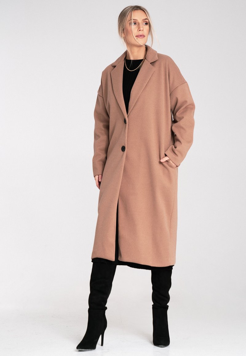 Brązowy płaszcz w kratę houndstooth z wykładanym kołnierzem, dużymi guzikami z przodu, kieszeniami bocznymi i średniej długości krojem. Noszony z czarnymi kozakiem za kolano.