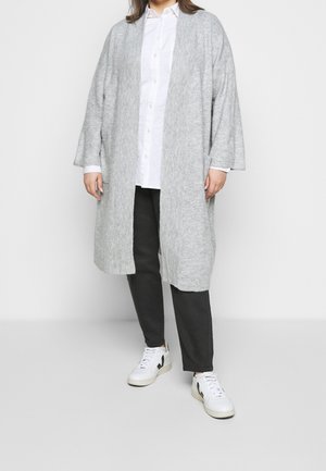 Personne portant un long cardigan gris clair, une chemise blanche à boutons, un pantalon noir et des baskets blanches, debout devant un fond uni.