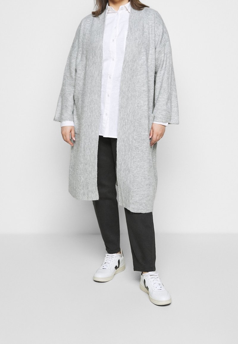 Personne portant un long cardigan gris clair, une chemise blanche à boutons, un pantalon noir et des baskets blanches, debout devant un fond uni.