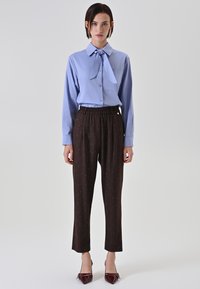 Femme portant un chemisier bleu à manches longues avec un col noué, un pantalon sombre à motifs taille haute et des escarpins pointus bordeaux, debout sur un fond blanc.
