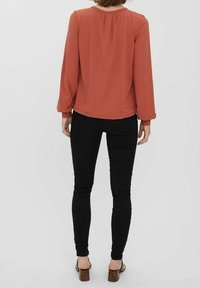 Blouse à manches longues couleur rouille, avec une coupe décontractée et un pli au dos, associée à un jean skinny noir et des sandales marron sur un fond neutre.
