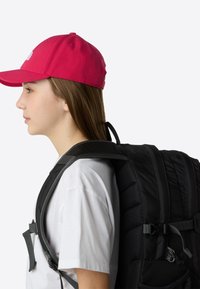 Svart ryggsäck med vadderade axelremmar och justerbara clips, bärs av en person i en vit skjorta och en ljusrosa keps, sedd från sidan.