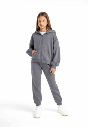 Szary hoodie na zamek z dużymi kieszeniami i ściągaczami na rękawach, zestawiony z pasującymi spodniami dresowymi. Wykonany z miękkiego materiału, ma luźny krój. Białe sneakersy.