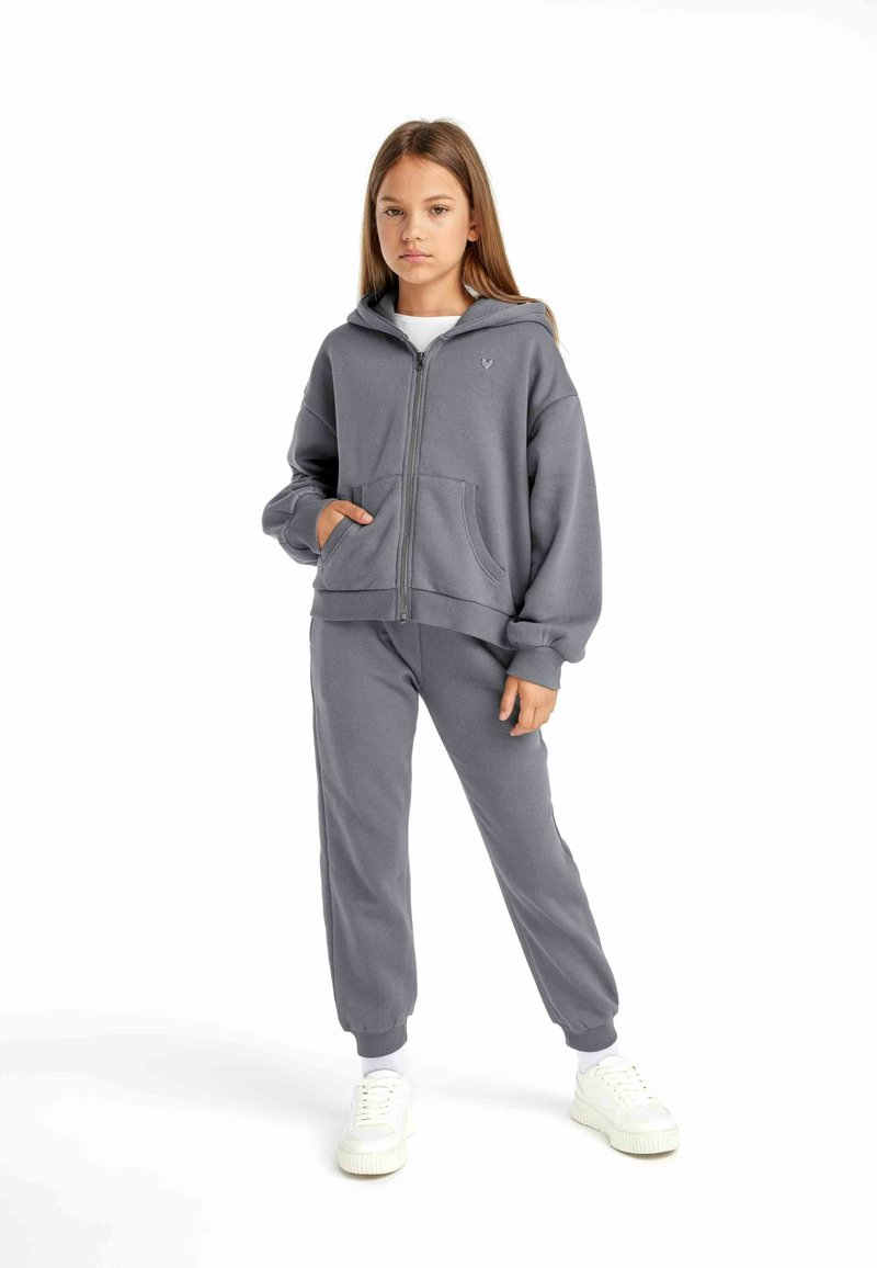 Sudadera gris con cremallera y grandes bolsillos, acompañada de pantalones de chándal a juego. Presenta un tejido suave y un corte relajado. Zapatillas blancas.