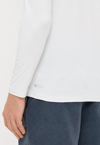 Λευκό πουκάμισο με μακριά μανίκια, κατασκευασμένο από ελαφρύ ύφασμα, το οποίο διαθέτει ετικέτα UPF 50+ στο τελείωμα. Η κοντινή λήψη αποτυπώνει λεπτομέρειες του μανικιού και από τη πλάγια όψη.