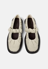 Scarpe beige con forma arrotondata, dotate di una tomaia in tessuto arricciato e suola in gomma nera, progettate per offrire una sensazione di morbidezza e ammortizzazione.