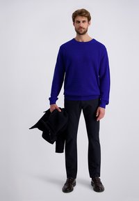 Koninklijk blauwe gebreide trui met een ronde hals, gecombineerd met donkere broeken. Het model houdt een zwart jack vast en draagt donkerbruine schoenen.