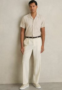 Camisa de manga corta beige con un bolsillo, combinada con pantalones crema y zapatillas de tono claro, acentuada por un cinturón marrón oscuro.
