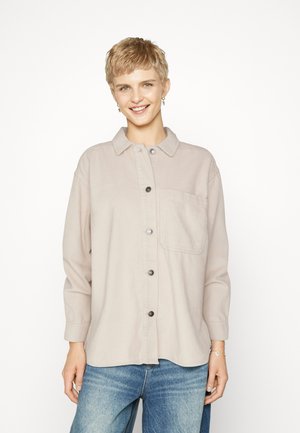 Chemise en velours côtelé beige clair avec des manches longues, boutonnière à l'avant, col et une seule poche poitrine. Portée avec un jean bleu.