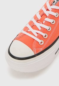 Orange canvas sneaker with white laces, rounded toe, and rubber sole. Features silver eyelets and black rubber trim on the base.