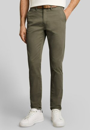 Mann trägt olivgrüne Slim-Fit-Hose, weiße Sneakers und einen braunen Gürtel, steht mit einer Hand in der Tasche vor schlichtem Hintergrund.