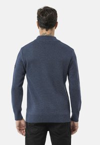 Pull en tricot bleu marine à manches longues, avec un col et un ourlet côtelés. Présente une texture subtile et une patch logo sur l'épaule gauche.