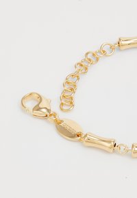 Bracelet en chaîne dorée avec fermoir à homard, maillons ajustables et une étiquette ovale gravée "GUESS" sur fond blanc.