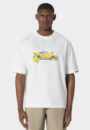 Giovane uomo con indosso una maglietta bianca con grafica di un'auto sportiva gialla e un limone, abbinata a pantaloni beige, in piedi davanti a uno sfondo neutro.