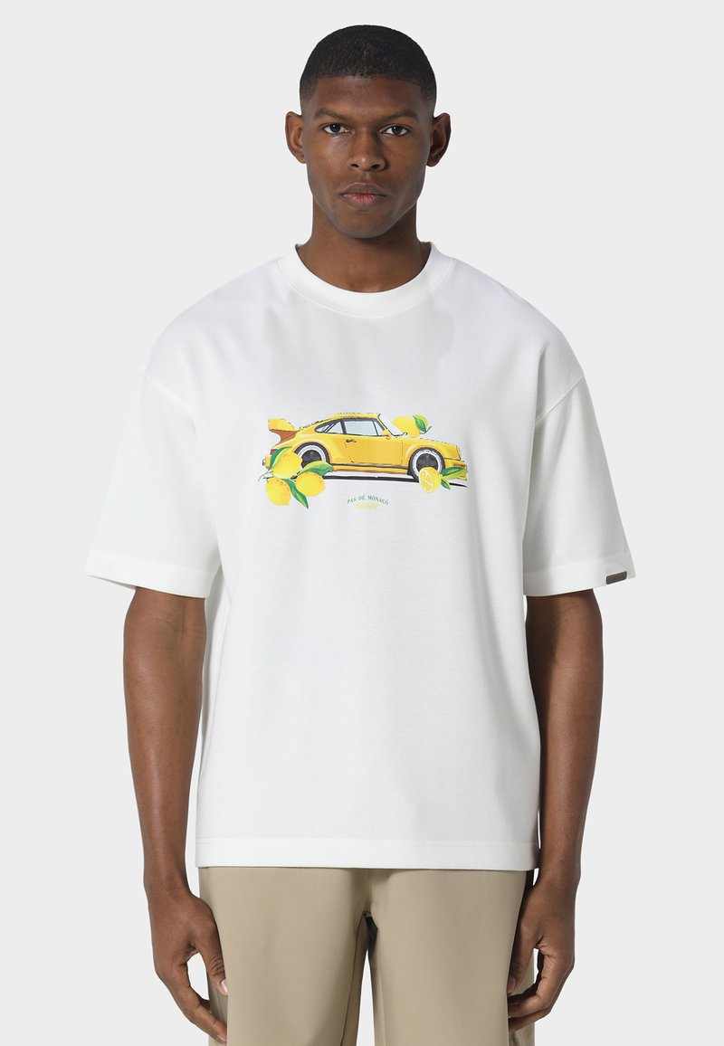 Giovane uomo con indosso una maglietta bianca con grafica di un'auto sportiva gialla e un limone, abbinata a pantaloni beige, in piedi davanti a uno sfondo neutro.