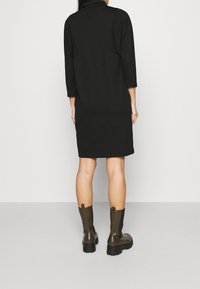 Czarna sukienka do kolan z trzema czwarciami rękawów i wysokim dekoltem. Noszona z brązowymi, dużymi butami na obcasie z elastycznymi panelami bocznymi.