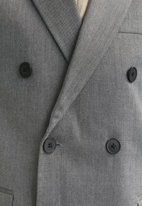 Gros plan sur un blazer gris croisé à boutons noirs avec une texture subtile à fines rayures, porté sur un vêtement en tricot beige.