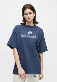 Νέος με κοντά καστανά μαλλιά, φορώντας ένα χαλαρό ναυτικό μπλε μπλουζάκι Nike Club και ανοιχτόχρωμο παντελόνι, μπροστά από μια απλή φόντο.