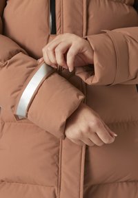 Chaqueta tipo puffer en un marrón cálido con un acento plateado brillante en el puño. Tela gruesa y texturizada con un diseño acolchado. Cierre negro visible.