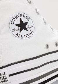 Baskets montantes en toile blanche avec un logo étoile noir, une semelle rayée noir et blanc, des œillets métalliques et une tige en tissu texturé.