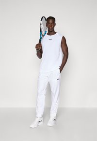 Λευκή αθλητική ενδυμασία με αμάνικο τοπ και στενό  παντελόνι, και τα δύο με λογότυπο. Άνδρας κρατάει ρακέτα τένις, φορώντας αθλητικά παπούτσια.