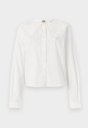 Camisa blanca de manga larga con un gran cuello de encaje, cierre frontal de botones y un dobladillo ligeramente cortado. Fabricada en tela ligera.