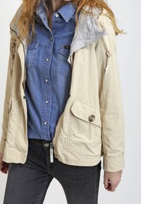 Beige letvægtsjakke med grå hætte, to forlommer og lynlåslukning, lagdelt over en blå denimskjorte.