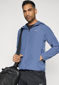 Ελαφρύ μπλε μπουφάν Nike με φερμουάρ μπροστά, κουκούλα και ελαστικούς καρπούς. Το μοντέλο κρατά μια μαύρη τσάντα, φορώντας μαύρη μπλούζα και σκούρα παντελόνια.