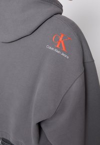 Szara bluza z kapturem z czerwonym logo "cK" i białym napisem "Calvin Klein Jeans" na górnej części pleców przy ramieniu.