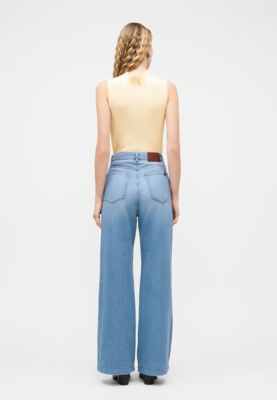 Femme aux longs cheveux blonds ondulés, portant un haut jaune sans manches et un jean bleu taille haute à jambes larges, debout de dos sur fond blanc.