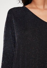 Black top with a V-neckline, featuring a textured fabric that shimmers with multicolored flecks throughout the surface.
