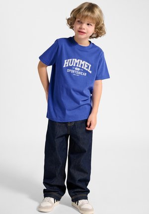 Ung dreng med lyst krøllet hår iført en blå Hummel sports t-shirt, mørke jeans og hvide sneakers, stående mod en ensfarvet baggrund.