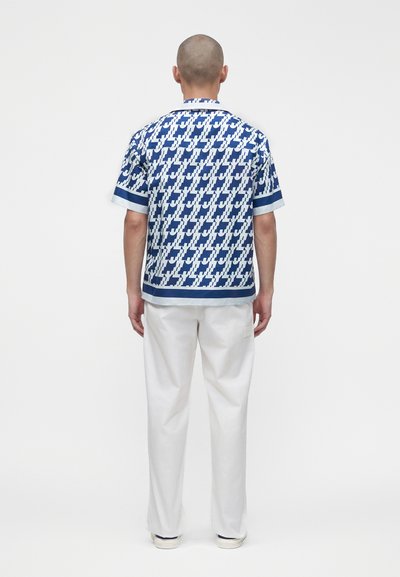 Hombre con la cabeza rapada que lleva una camisa de manga corta con estampado azul y blanco y pantalones blancos, de espaldas sobre un fondo blanco.