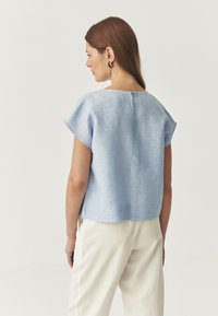 Lyseblå linned blouse med korte ærmer og rund hals, med knapdetalje på ryggen og en afslappet pasform. Båret sammen med cremefarvede bukser.
