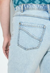 Vaaleansiniset farkut, joissa on korkea vyötärö, takatasku, jossa on kaareva ommel, sileä pinta ja minimaalinen varustelu.