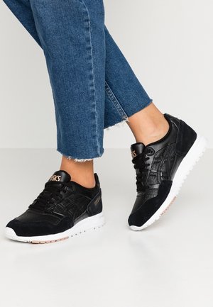 Czarne teksturowane buty ASICS noszone z krótkimi niebieskimi dżinsami na jasnoszarym tle podłogi.