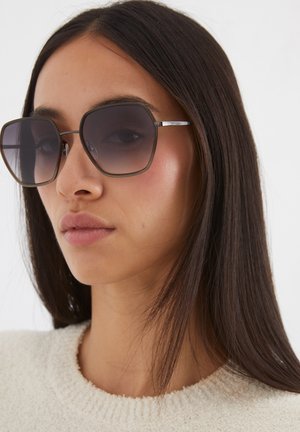 Bruine oversized zonnebril met een vierkant montuur, gradient lenzen en dunne zilveren metalen accenten. Het haar is glad en steil.